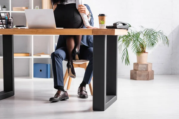 Partial view of secretary sitting on desk near laptop and seducing colleague in office — Stock Photo