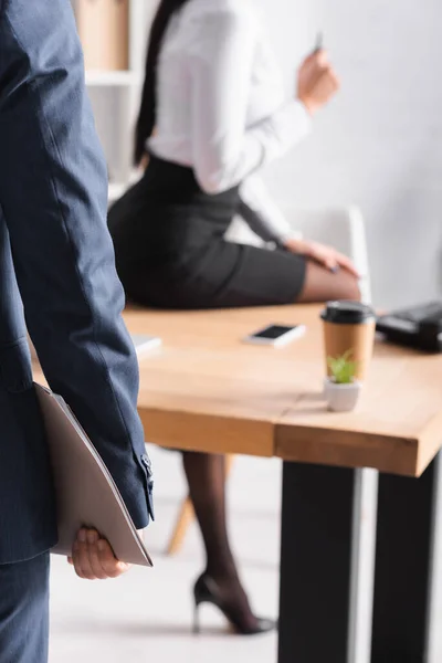 Частковий вид на бізнесмена з паперовою текою біля сексуальної бізнес-леді, що сидить на столі на розмитому фоні — стокове фото