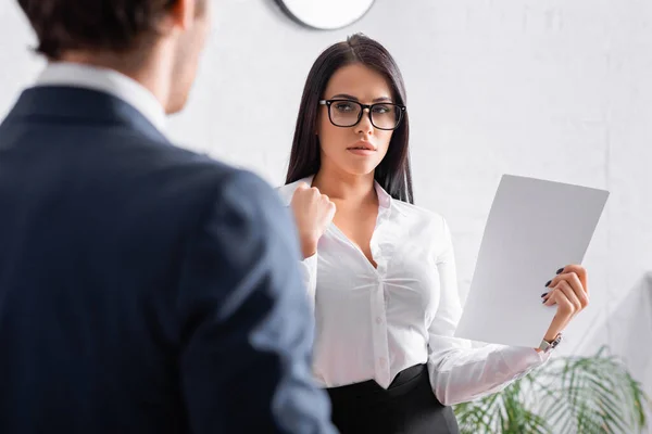 Verführerische brünette Sekretärin hält Papier in der Hand, während sie mit einem Geschäftsmann im verschwommenen Vordergrund flirtet — Stockfoto