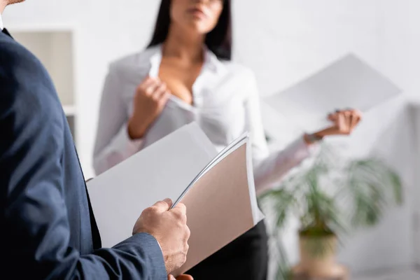 Vista parcial de hombre de negocios con carpeta de papel cerca de mujer de negocios seductora mostrando busto en la oficina, fondo borroso - foto de stock