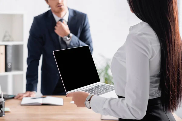 Vue recadrée d'une femme d'affaires sexy tenant un ordinateur portable avec écran vide près d'un collègue ajustant la cravate sur fond flou — Photo de stock