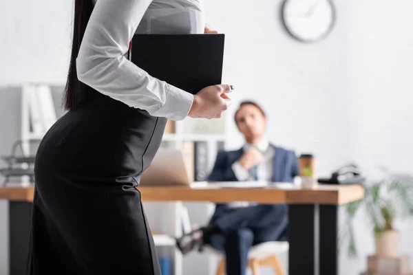 Sexy Sekretärin mit Notizbuch in der Nähe Geschäftsmann sitzt am Arbeitsplatz auf verschwommenem Hintergrund — Stockfoto