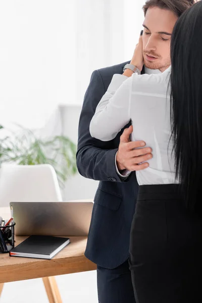 Vista trasera de hombre de negocios con los ojos cerrados abrazando secretaria morena en la oficina - foto de stock