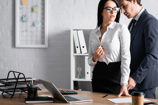 Young businessman near passionate secretary unbuttoning blouse near desk, blurred foreground — Stock Photo