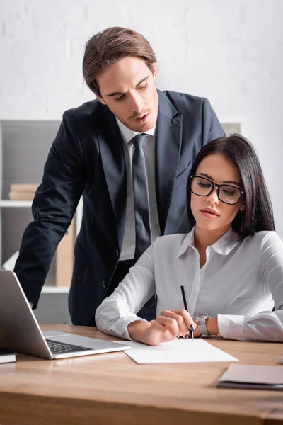 Uomo d'affari vicino sensuale bruna segretario seduto sul posto di lavoro vicino al computer portatile — Foto stock