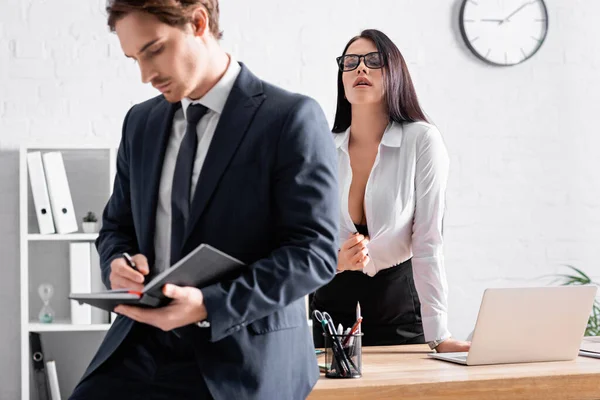 Apasionada mujer de negocios desabotonar blusa cerca colega escribir en cuaderno en primer plano borrosa - foto de stock