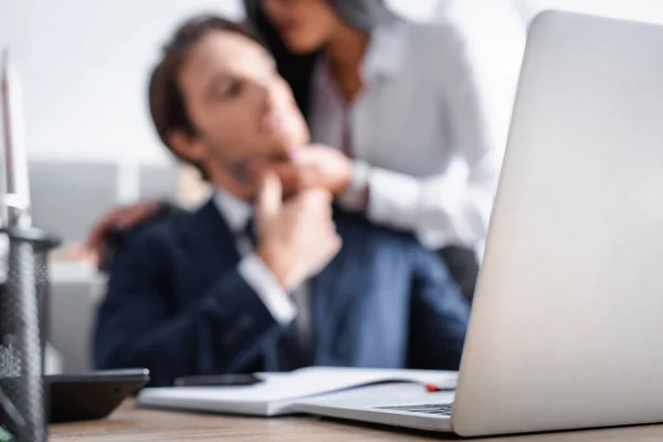 Selective focus of laptop near businesswoman flirting with young colleague on blurred background — Stock Photo