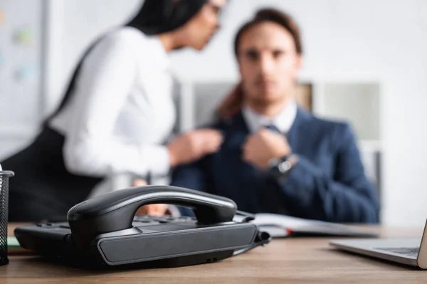 Selektiver Fokus des Festnetztelefons in der Nähe einer Sekretärin, die mit einem Geschäftsmann flirtet, auf verschwommenem Hintergrund — Stockfoto