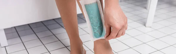 Cropped view of woman removing wax stripe on leg in bathroom, banner — Stock Photo