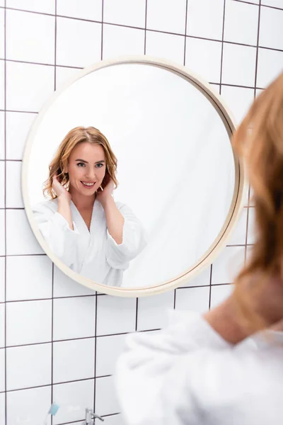 Счастливая женщина в халате регулирует волосы, глядя на зеркало в ванной комнате — стоковое фото