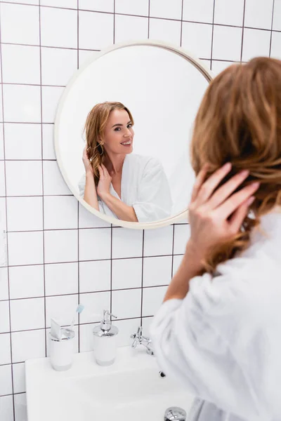 Mujer alegre en albornoz ajustando el cabello mientras mira el espejo en el baño - foto de stock
