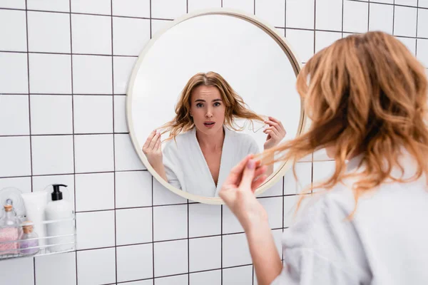 Femme mécontente en peignoir ajuster les cheveux près du miroir dans la salle de bain — Photo de stock