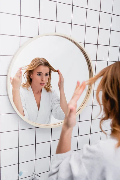 Displeased woman in bathrobe adjusting hair while looking at mirror in bathroom — Stock Photo