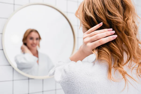 Donna in accappatoio regolazione capelli vicino specchio su sfondo sfocato — Foto stock