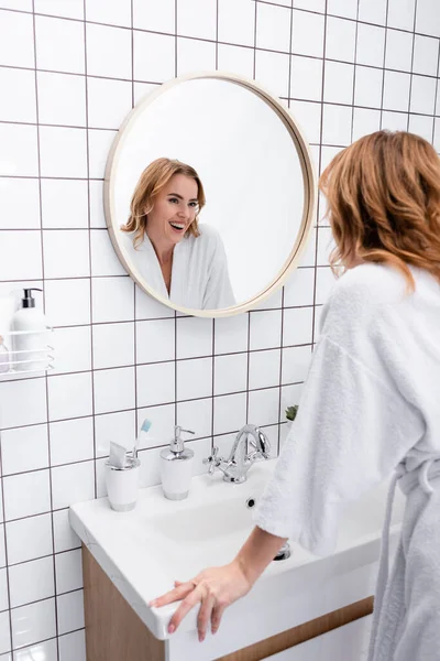 Fröhliche Frau im Bademantel lächelt, während sie im Badezimmer in den Spiegel schaut — Stockfoto