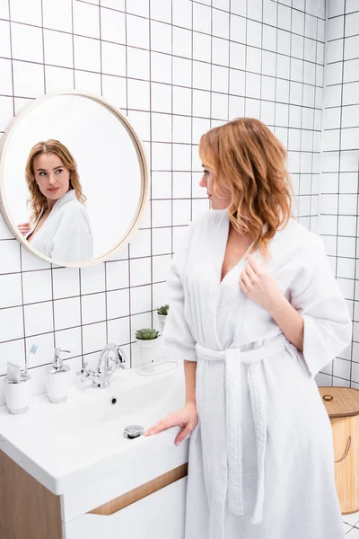 Женщина в белом халате смотрит в зеркало в ванной комнате — стоковое фото