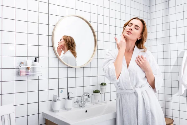 Женщина с закрытыми глазами наносит крем для лица в ванной комнате — стоковое фото