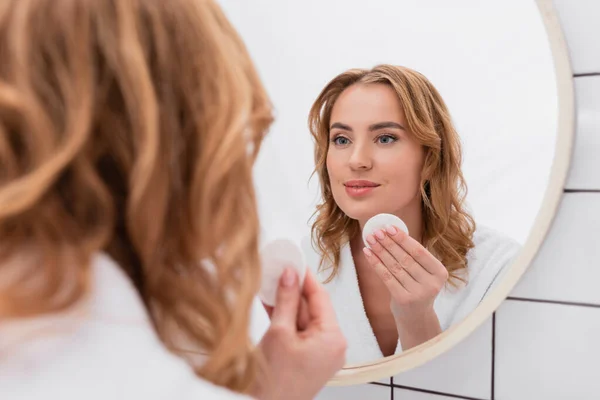 Mulher feliz aplicando toner com almofada de algodão no rosto e olhando para o espelho — Fotografia de Stock