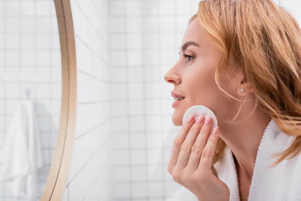Side view of woman applying toner with cotton pad on face and looking at mirror — Stock Photo