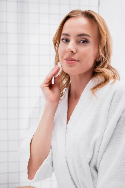 Femme souriante appliquant toner avec coton sur le visage et en regardant la caméra — Photo de stock