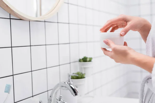 Vista recortada de la mujer sosteniendo frasco con crema facial cerca del grifo en el baño - foto de stock