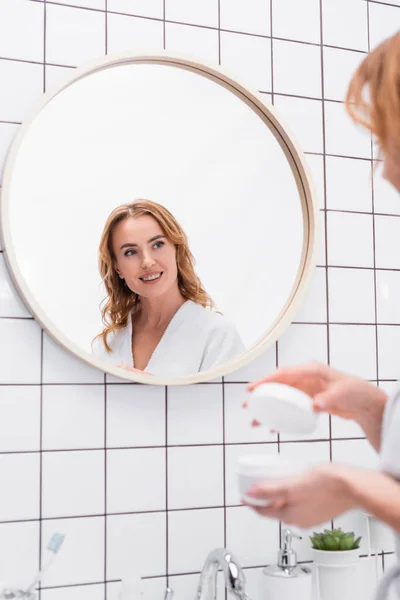 Mujer alegre sosteniendo tarro con crema facial y mirando el espejo en el baño - foto de stock