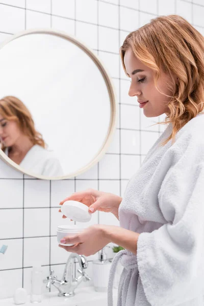 Mujer alegre mirando frasco con crema facial en el baño - foto de stock