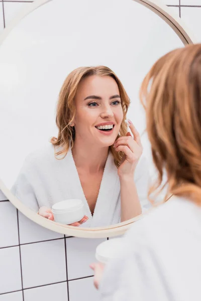 Mulher feliz segurando frasco e aplicando creme facial enquanto olha para o espelho — Fotografia de Stock