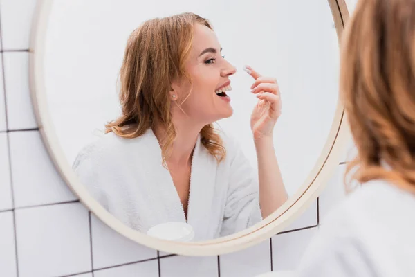 Mulher sorrindo segurando frasco e aplicando creme facial enquanto olha para o espelho — Fotografia de Stock