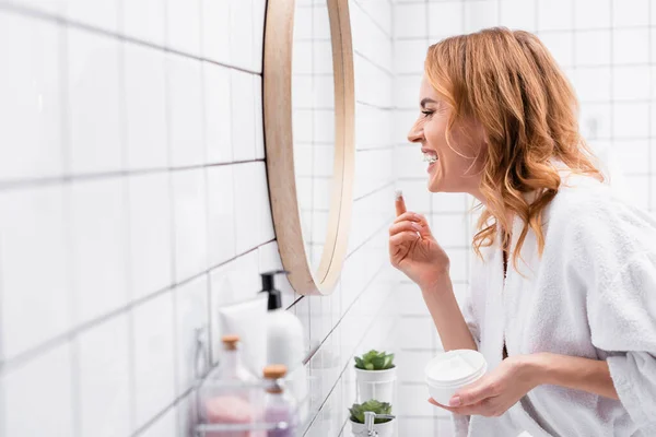 Visão lateral da mulher sorridente segurando frasco e aplicando creme facial enquanto olha para o espelho perto de garrafas em primeiro plano borrado — Fotografia de Stock