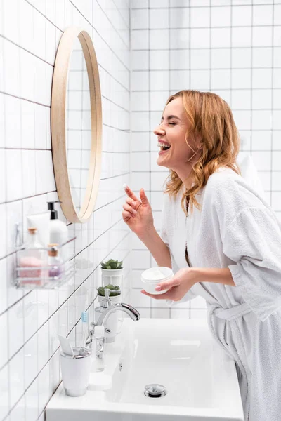 Sonriente mujer sosteniendo frasco con crema facial y riendo cerca del espejo y botellas en primer plano borrosa - foto de stock