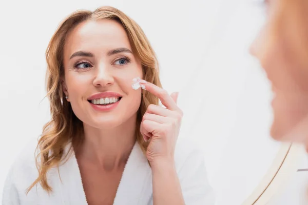 Reflet de femme souriante appliquer crème cosmétique sur le visage dans la salle de bain — Photo de stock