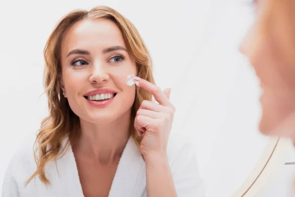 Reflejo de la mujer sonriente aplicando crema cosmética en la cara en el baño - foto de stock