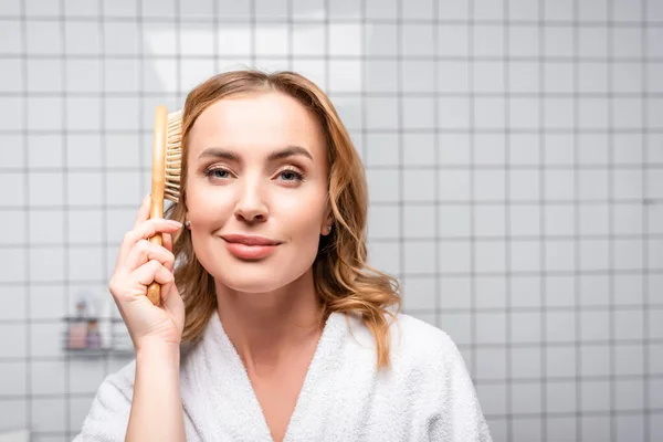 Zufriedene Frau im weißen Bademantel beim Haareputzen im Badezimmer — Stockfoto