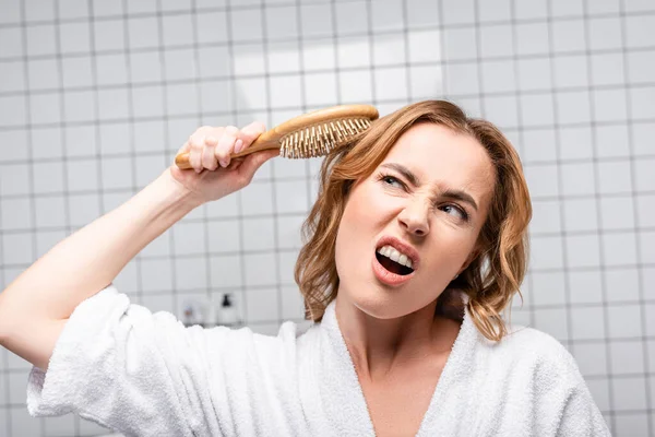 Mujer disgustada en albornoz blanco cepillarse el pelo en el baño - foto de stock