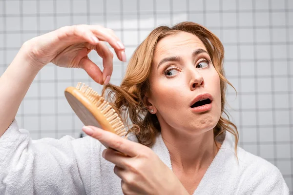 Mujer insatisfecha en albornoz cepillarse el pelo en el baño - foto de stock