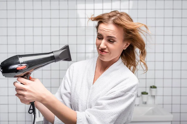 Unzufriedene Frau im weißen Bademantel trocknet glänzendes Haar im Badezimmer — Stockfoto