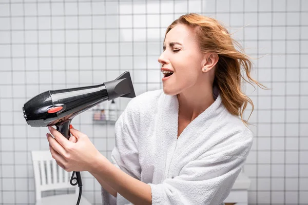 Woman in white bathrobe drying shiny hair and singing in bathroom — Stock Photo