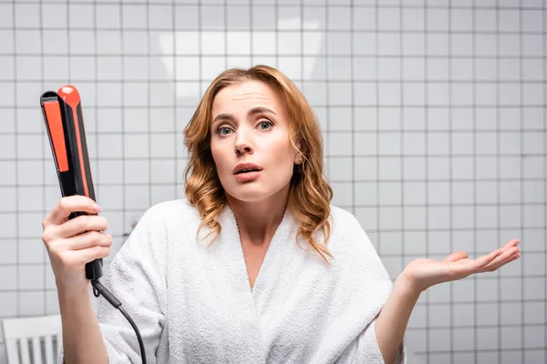 Confused woman in white bathrobe holding hair straightener in bathroom — Stock Photo