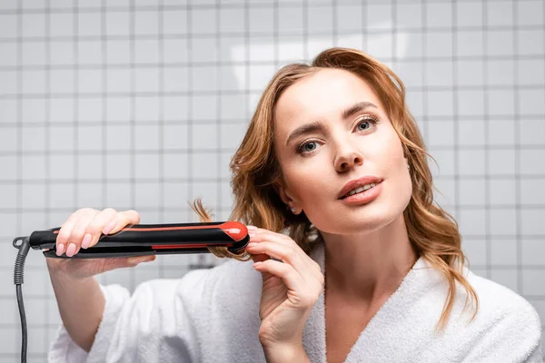 Frau im weißen Bademantel benutzt Haarglätter im Badezimmer — Stockfoto