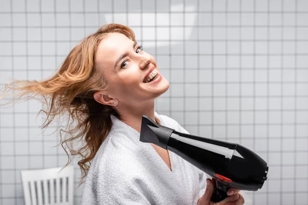 Femme joyeuse en peignoir sèche-cheveux dans la salle de bain — Photo de stock