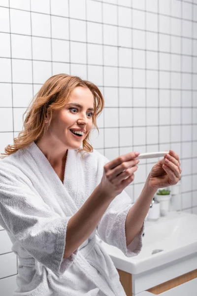 Happy woman in white bathrobe holding pregnancy test in bathroom — Stock Photo