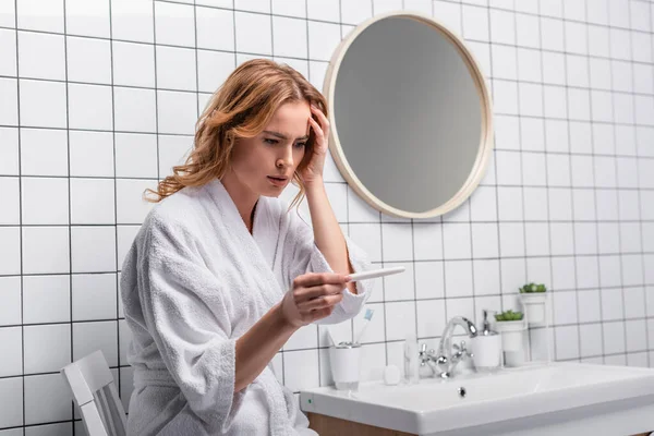 Femme inquiète en peignoir blanc tenant test de grossesse dans la salle de bain — Photo de stock