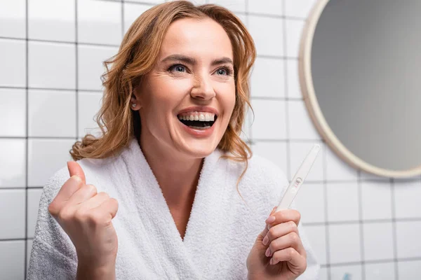 Aufgeregte Frau im weißen Bademantel hält Schwangerschaftstest im Badezimmer — Stockfoto
