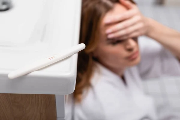Test di gravidanza con esito positivo vicino a donna preoccupata su sfondo sfocato in bagno — Foto stock