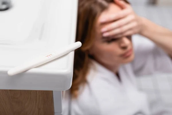 Pregnancy test with positive result near worried woman on blurred background in bathroom — Stock Photo