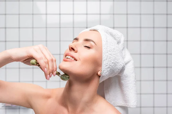 Mujer feliz con los ojos cerrados y toalla en la cabeza usando rodillo de jade en el baño - foto de stock