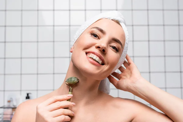 Mujer alegre con toalla en la cabeza usando rodillo de jade en el baño — Stock Photo