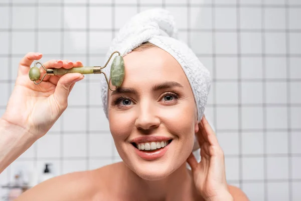 Happy woman with towel on head using jade roller in bathroom — Stock Photo