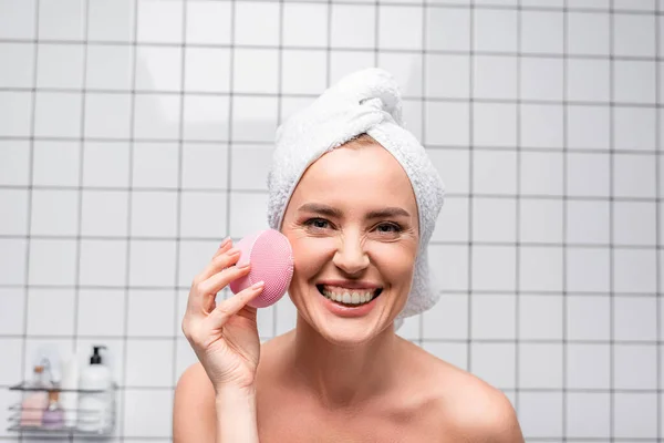 Mulher alegre em toalha na cabeça segurando escova de silicone de limpeza no banheiro — Fotografia de Stock